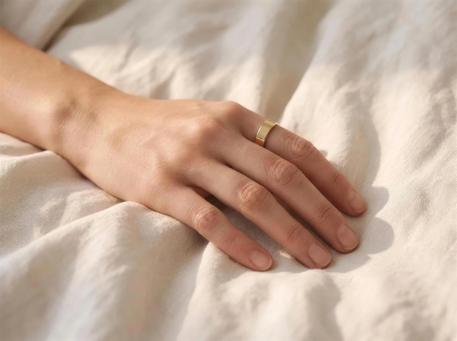 A hand at rest on linen bedding at dawn, wearing the gold Vexo ring